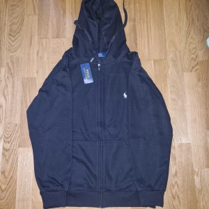 Svart hoodie från Polo Ralph Lauren - Svart hoodie från Polo Ralph Lauren med dragkedja framtill och klassisk huva. Tröjan har två fickor och den ikoniska vita logon broderad på bröstet. Perfekt för en avslappnad och stilren look.