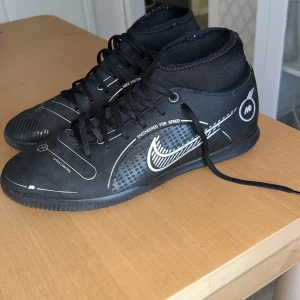 Svarta Nike fotbollsskor med snörning - Säljer ett par svarta Nike fotbollsskor med vita detaljer och tryck. Skorna har snörning, rund tå och är tillverkade i syntetmaterial. De har en tight passform och är designade för inomhusspel. Snygg och sportig look med Nike-logga på sidan.
