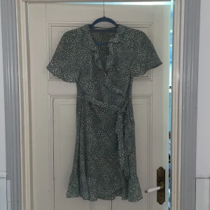 Grönprickig omlottklänning med volang - Söt kort grön klänning med vita prickar, omlottmodell och knytband i midjan. Klänningen har korta ärmar, volangkant nertill och en lätt, flowy känsla. Perfekt för dig som gillar lekfulla mönster och en avslappnad men trendig stil.