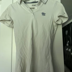 Vit piké t-shirt från Hilfiger Denim - Vit piké t-shirt från Hilfiger Denim med klassisk krage och knappar framtill. T-shirten har korta ärmar och en liten broderad logga på bröstet. Perfekt för en clean och stilren look.