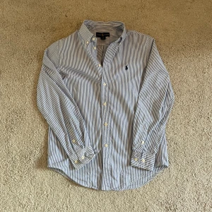 Randig skjorta från Ralph Lauren - Klassisk skjorta från Ralph Lauren i blåvitt randigt mönster. Skjortan har lång ärm, knappar framtill och broderad logga på bröstet. Tillverkad i mjuk bomull och passar perfekt till en stilren look. Obs hr en fläck som inte går bort på arm bågen.