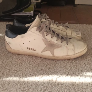 Golden Goose Superstar sneakers vit/grå - Golden Goose Superstar sneakers i vit skinn med grå mockadetaljer och grå snörning. Ikonisk stjärna på sidan och svart hälparti med logga. Platt sula och rund tå. Skejtat i dessa skor vilket är sorligt, fråga inte varför🫨 annars är de i super bra kondition!