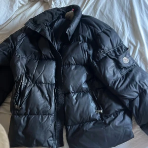 Svart pufferjacka från Moncler - Säljer en svart pufferjacka från Moncler med hög krage och två dragkedjeförsedda fickor. Jackan har klassisk Moncler-logga på ärmen och quiltad design som ger en riktigt fet vinterlook. Perfekt för kalla dagar när du vill hålla stilen.