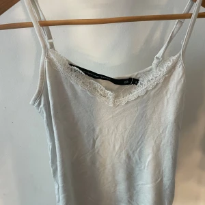 Vit linnetopp med spets från 157 - Säljer en vit linnetopp från 157 med tunna axelband och v-ringning dekorerad med spets. Toppen är enkel och stilren, perfekt att matcha med jeans eller kjol. Materialet känns mjukt och lätt mot huden. Använd 2 gånger