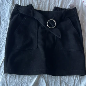 Svart kortkjol med bälte och fickor - Svart kortkjol med bredt bälte och silverfärgat spänne i midjan. Kjolen har två stora fickor framtill och en diskret dragkedja bak. Snygg och enkel design som passar till många olika stilar.