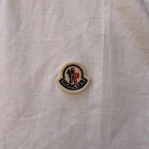 Vit Moncler t-shirt med logga - Snygg vit t-shirt från Moncler med klassisk rund hals och broderad logga på bröstet. Tillverkad i mjuk bomull och är i storlek S-M. Oanvänt skick och fraktas inom 24h✅