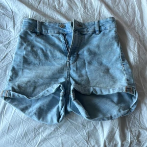Ljusblå jeansshorts med uppvik - Säljer ett par ljusblå jeansshorts med klassisk femficksdesign och uppvikta benslut. Shortsen har bälteshällor, knapp och dragkedja framtill samt två bakfickor. Perfekta för varma dagar och enkel att matcha med allt.
