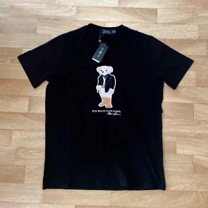 Helt ny Svart Polo Bear t-shirt från Ralph Lauren - Svart t-shirt från Ralph Lauren med ikoniska Polo Bear broderad framtill. Klassisk passform med rund hals och korta ärmar. Tillverkad i mjuk bomull som känns skön mot huden. Perfekt statement-plagg för dig som gillar stilrena och coola detaljer.