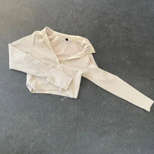 Beige ribbad croppad hoodie - Säljer en beige croppad hoodie med ribbad struktur och långa ärmar. Tröjan har en huva och är tillverkad i ett mjukt, stretchigt material som sitter snyggt och bekvämt. Perfekt för en chill och trendig look.