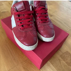 Valentino Garavani röda sneakers - Snygga röda och vita sneakers från Valentino Garavani med klassiska nitar på sulan. Skorna har röd ovandel i skinn, vita detaljer på sidor och häl samt röda skosnören. Perfekta för dig som vill sticka ut med en lyxig streetstyle-vibe.