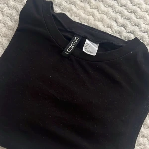 Svart genomskinlig t-shirt från DIVIDED - En svart, klassisk t-shirt från DIVIDED by H&M i storlek S. T-shirten har rund hals och korta ärmar, perfekt för en clean och enkel look. Tröjan är genomskinlig , fint att ha med ett linne eller liknande under.