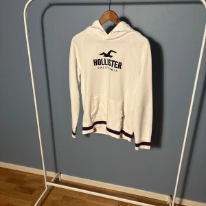 Vit hoodie från Hollister XS - Vit hoodie från Hollister med broderad logga och text på bröstet. Tröjan har huva, känguruficka och ribbade muddar med marinblå och röda ränder. Perfekt för chill dagar och snygg till jeans eller leggings.
