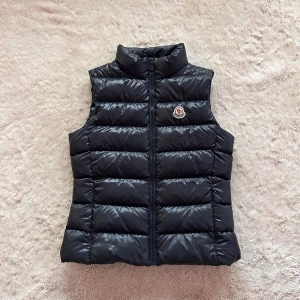 Marinblå dunväst från Moncler - Snygg marinblå dunväst från Moncler med quiltad design och hög krage. Västen har dragkedja framtill och ett broderat Moncler-märke på bröstet. Skick: 8/10 (endast använd fåtal gånger) Stolek: 14Y/164 (passar dig som använder XS vandligtvis) 