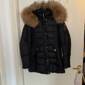 Svart dunjacka Rockandblue med päls - Snygg svart dunjacka från Rockandblue med stor fluffig päls runt huvan. Jackan har quiltade detaljer, dragkedjor på fickorna och Rockandblue-logga på ärmen. Perfekt för kalla dagar när du vill hålla stilen och värmen.