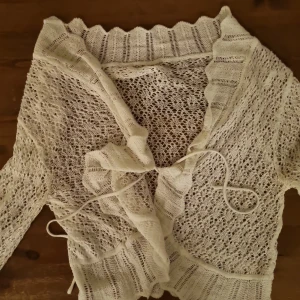 Vit stickad kofta med knyt - Supersöt vit stickad kofta med knyt framtill och vågig kant vid halsen. Koftan är tunn, har ett genombrutet mönster och 3/4 ärmar. Perfekt att slänga över en topp för en chill och romantisk look. Storlek Okänd men gissar på storlek S