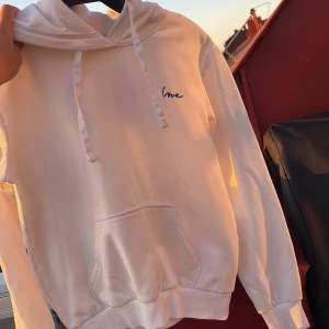 Vit hoodie med broderad text - Vit hoodie med broderad svart text 'love' på bröstet. Klassisk känguruficka framtill, snörning i huvan och ribbade muddar vid ärmslut och midja. Perfekt för chill dagar och enkel att matcha med jeans eller mjukisbyxor.