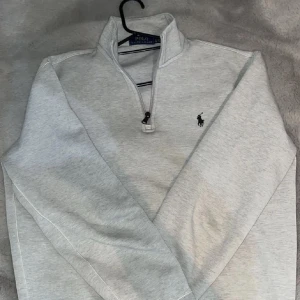 Grå half zip tröja från Polo Ralph Lauren - Snygg ljusgrå långärmad tröja från Polo Ralph Lauren med klassisk broderad logga på bröstet. Tröjan har en half zip-dragkedja och hög krage, perfekt för lager-på-lager. Mjuk bomullsblandning som är skön att bära hela dagen. Tveka inte på att fråga på 