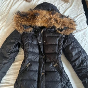 Svart dunjacka Rockandblue  - Säljer en svart dunjacka från Rockandblue med stor huva och snygg päls i bruna toner. Jackan har dragkedjor på fickorna, markerad midja och Rockandblue-logga på ärmen. Perfekt för kalla dagar och riktigt stilren look, original pris ligger runt 2000-2500 kr. Som man ser på sista bilden så är den trasig vid luvan men märks ej av och påverkar inte jackan på något sätt 😊