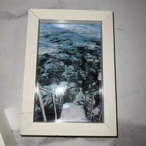 Fin tavla  - Bilderna visar en vit fotoram med ett foto av en trappa ner i vatten. Ramen har ett stöd för att stå på bord och kan även hängas på vägg. Det finns en spricka och ett litet skadat hörn på sidan