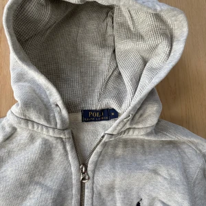 Grå hoodie från Polo Ralph Lauren - Snygg grå hoodie från Polo Ralph Lauren med dragkedja framtill och klassisk huva. Tröjan har långa ärmar, fickor på magen och den ikoniska broderade loggan på bröstet. Perfekt för en chill och stilren look.
