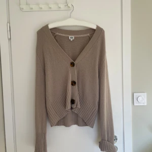 Beige kofta med stora knappar Twist & Tango - Säljer en beige stickad kofta från Twist & Tango med stora bruna knappar framtill. Koftan har v-ringning, ribbade muddar och en croppad passform som ger en trendig look. Perfekt för lager-på-lager under hösten och våren.