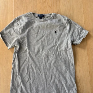 Grå t-shirt från Polo Ralph Lauren - Klassisk grå t-shirt från Polo Ralph Lauren med rund halsringning och broderad logga på bröstet. Tillverkad i mjuk bomull och har en enkel, stilren design som passar till allt. Perfekt för dig som gillar en clean och tidlös look.