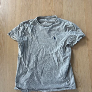 Grå t-shirt från Polo Ralph Lauren - Snygg grå t-shirt från Polo Ralph Lauren med klassisk rund hals och broderad marinblå logga på bröstet. Tillverkad i mjuk bomull och har en enkel, stilren design som passar till det mesta.