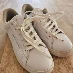 Vita sneakers från Tommy Hilfiger - Snygga vita sneakers från Tommy Hilfiger med klassisk design. Skorna har rund tå, platt sula och är tillverkade i skinn med vita snören. Diskret logga på sidan och tungan. Perfekta för en clean och stilren look. Mycket lite använda. Vitare i verkligheten än på bilderna.