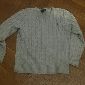 Grå kabelstickad tröja Polo Ralph Lauren - Kabelstickad grå tröja från Polo Ralph Lauren med klassisk rund hals och röd broderad logga på bröstet. Tröjan har ribbade muddar och är tillverkad i mjuk bomull, perfekt för lager-på-lager under höst och vinter.
