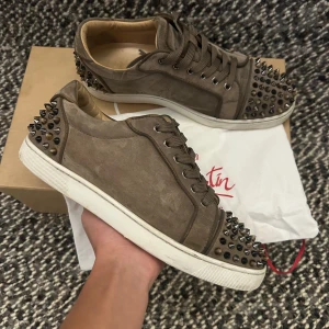 Bruna mockasneakers med nitar Louboutin - Säljer ett par bruna sneakers från Christian Louboutin i mjuk mocka med coola nitar i svart, silver och brons på tå och häl. Skorna har vita platta sulor och snörning framtill. Perfekta för dig som vill sticka ut med en edgy vibe.