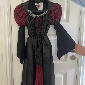 Vampyrklänning 110 cm lång/passar till 5-8 år - Cool HALLOWEEN klänning passar till 5-8 år, med puffärmar och snörning framtill. Klänningen har röda detaljer med spindelnätsmönster och trumpet-ärmar. Perfekt för halloween eller temafest när du vill sticka ut med en dramatisk look. Sömmen i kragen har spruckit lite(lätt att laga) , annars helt i nyskick. Nypris 750 kr.