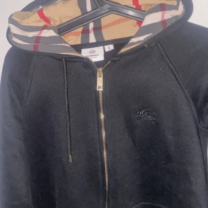 Svart Burberry hoodie med dragkedja - Svart hoodie från Burberry med klassiskt rutigt foder i huvan, guldfärgad dragkedja och broderad logga på bröstet. Tröjan har snörning i huvan och två fickor framtill. Perfekt för dig som vill ha en stilren och lyxig look. Skulle säga att den passar för dig som har S bättre.