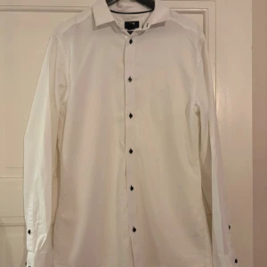 Vit skjorta med svarta knappar - Klassisk vit skjorta från H&M med svarta knappar som ger en snygg kontrast. Skjortan har lång ärm, normal passform och är tillverkad i bomull. Perfekt för dig som gillar stilrena och enkla plagg med en twist.