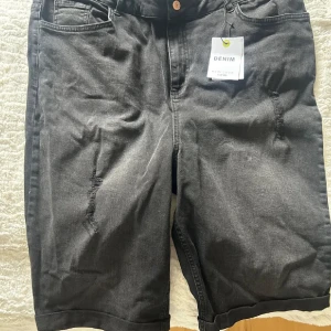 Svarta denimshorts  - Säljer ett par svarta denimshorts De har klassisk femficksdesign, råa slitningar framtill och på bakfickorna samt knapp och dragkedja i gylfen. Perfekta för en avslappnad och cool stil.