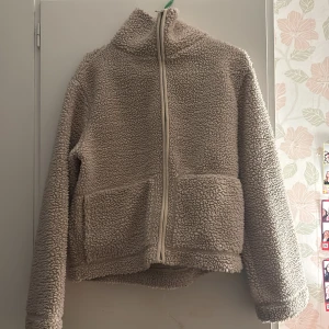 Beige teddyjacka med dragkedja - Mysig beige teddyjacka med hög krage och hel dragkedja framtill. Jackan har två stora fickor på framsidan och är tillverkad i fluffigt fleece-material. Perfekt för kalla dagar när du vill vara både varm och stilren.