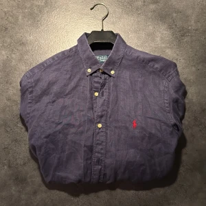 Blå skjorta från Polo Ralph Lauren - Snygg marinblå skjorta från Polo Ralph Lauren med klassisk button-down krage och röd broderad logga på bröstet. Skjortan är i bomull och har slim fit-passform med långa ärmar och knappar framtill. Perfekt för en clean och stilren look. S men passar M