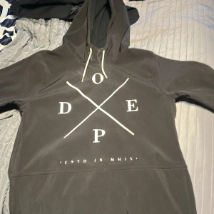 Svart hoodie från Dope med tryck - Svart hoodie från Dope med stort vitt tryck på bröstet och snörning i huvan. Tröjan har dragkedjor i sidorna och en klassisk känguruficka framtill. Perfekt för dig som gillar skidor eller snowboard 