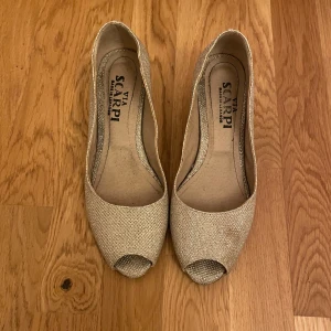 Glittriga pumps från Via Scarpi - Snygga pumps i guldigt glittrigt material från Via Scarpi. Skorna har öppen tå, låg klack och rundad form. Perfekta för dig som vill sticka ut med lite extra bling. Bekväma och stilrena med en elegant touch.