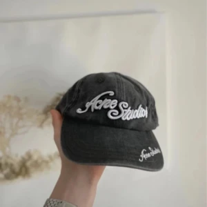 Acne Studios keps - Snygg grå keps från Acne Studios med vit broderad logga framtill och på skärmen. Justerbar rem baktill för perfekt passform. Klassisk och stilren design som passar till många outfits.