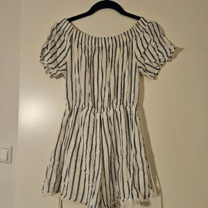 Randig kort klänning från Bershka - Superfin kort jumpsuit från Bershka i vitt med blåa ränder. Offshoulder-modell med puffiga korta ärmar och resår i midjan. Klänningen har knytband i midjan och spetsdetaljer längst ner. Köpt i Frankrike 