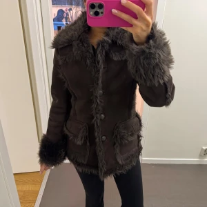 Brun jacka med fuskpälsdetaljer - Säljer en brun pälsjacka med fluffiga fuskpälsdetaljer på krage, ärmslut, fickor och längs kanterna. Jackan har stora knappar framtill och två fickor. Perfekt skick 