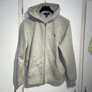 Grå hoodie från Polo Ralph Lauren  - Snygg ljusgrå hoodie från Polo Ralph Lauren i storlek M. Barn storlek. Passar 175-185. Tröjan har dragkedja, huva och två fickor framtill. Klassisk broderad logga på bröstet. Perfekt för chill dagar eller när du vill ha en clean look. Tröjan har en lite fläck i luvan som går bort med medel. Priset är ej hugget i sten✌🏽