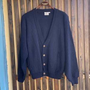 Mörkblå cardigan från Florentino 100% kvalité  - Klassisk mörkblå cardigan från Florentino med v-ringning och träknappar. Plagget är stickad i ett mjukt material och har två fickor framtill samt ribbade muddar vid ärmslut och nederkant. Perfekt under kyliga dagar när man ändå vill klä upp sig med möjligtvis en skjorta under. Storlek L är den sanna storleken. 