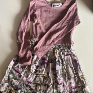 Rosa långärmad klänning från Molo - Supersöt klänning från Molo med mjukt rosa överdel och långärmad design. Kjolen har ett magiskt svampmönster i lila, beige och gröna toner. Tillverkad i bomull med lite elastan för extra komfort. Perfekt för dig som gillar unika prints och sköna material.