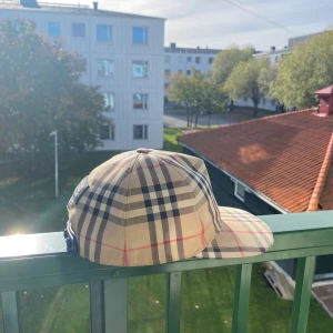 Burberry rutig keps med klassiskt mönster - Säljer en ikonisk keps från Burberry med det klassiska rutiga mönstret i beige, svart, rött och vitt. Kepsen har justerbar snapback och en svart patch med 'Burberry London England' bak. Perfekt accessoar för att lyfta din streetstyle.