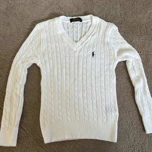 Vit kabelstickad tröja från Ralph Lauren - Vit kabelstickad tröja från Ralph Lauren med v-ringning och broderad logga på bröstet. Tröjan har lång ärm, ribbade muddar och sitter snyggt figurnära. Perfekt till jeans eller kjol för en klassisk look. 