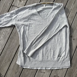 Ljusgrå v-ringad tröja från Esprit - Ljusgrå v-ringad tröja från Esprit i mjuk stickad bomullsblandning. Tröjan har långa ärmar, ribbade muddar och nederkant samt en diskret metallogo vid höften. Perfekt för lager-på-lager och chill stil.