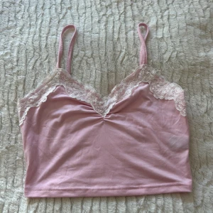 Ljusrosa linnetopp med spets - Supersöt ljusrosa linnetopp med tunna axelband och vit spetsdetalj längs v-ringad urringning. Toppen är croppad och har en mjuk, stretchig känsla. Perfekt att styla med jeans eller kjol för en trendig look.