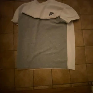 Nike t-shirt i vitt och grått - Nike t-shirt med vit överdel och grå nederdel, svart Nike-logga på bröstet. Klassisk rund hals och korta ärmar. Perfekt för en sportig och avslappnad stil.
