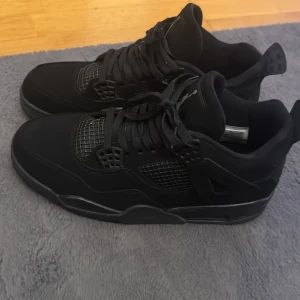 Svarta Nike Air Jordan 4 sneakers - Säljer ett par helsvarta Nike Air Jordan 4 sneakers med klassisk meshpanel, snörning och Jumpman-logga på plösen och sulan. Skorna har en chunky siluett och är tillverkade i en mix av skinn och syntetmaterial. Perfekta för dig som gillar streetwear och vill sticka ut med en ikonisk modell. Boxen till skorna följer med